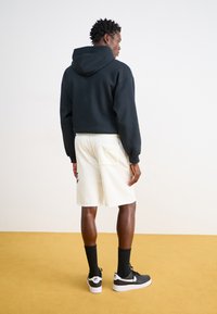 Svart huvtröja, krämfärgade shorts, svarta strumpor och svarta sneakers. Shorts har en ficka och en logotypdesign på sidan.