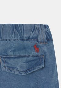 Pas modrých džínů s elastickým lemem a poutky na opasek, s zadní kapsou a malým červeným vyšitým logem koně nad kapsou.