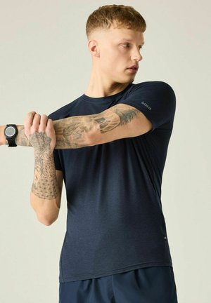 Młody mężczyzna z tatuażami rozciągający rękę, ubrany w ciemnoniebieską sportową koszulkę i smartwatch na jednolitym jasnym tle.