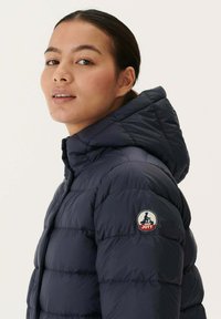 Marineblauwe gewatteerde jas met een capuchon, voorzien van gestikte segmenten. Inclusief een logo op de linkermouw. Gladde textuur en lichtgewicht ontwerp.