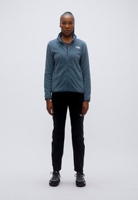 Τζάκετ από φλις σε αποχρώσεις γαλάζιου σχιστόλιθου με κλείσιμο με φερμουάρ· συνδυασμένο με μαύρο παντελόνι και μαύρα παπούτσια πεζοπορίας. Το μοντέλο στέκεται μπροστά σε απλό φόντο.