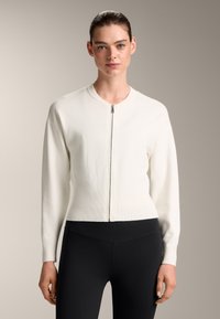 Bela zapinjalna jopica z dolgimi rokavi in rebrasto teksturo, z okroglim ovratnikom in tesnim pasom, v kombinaciji s črnimi leginami.