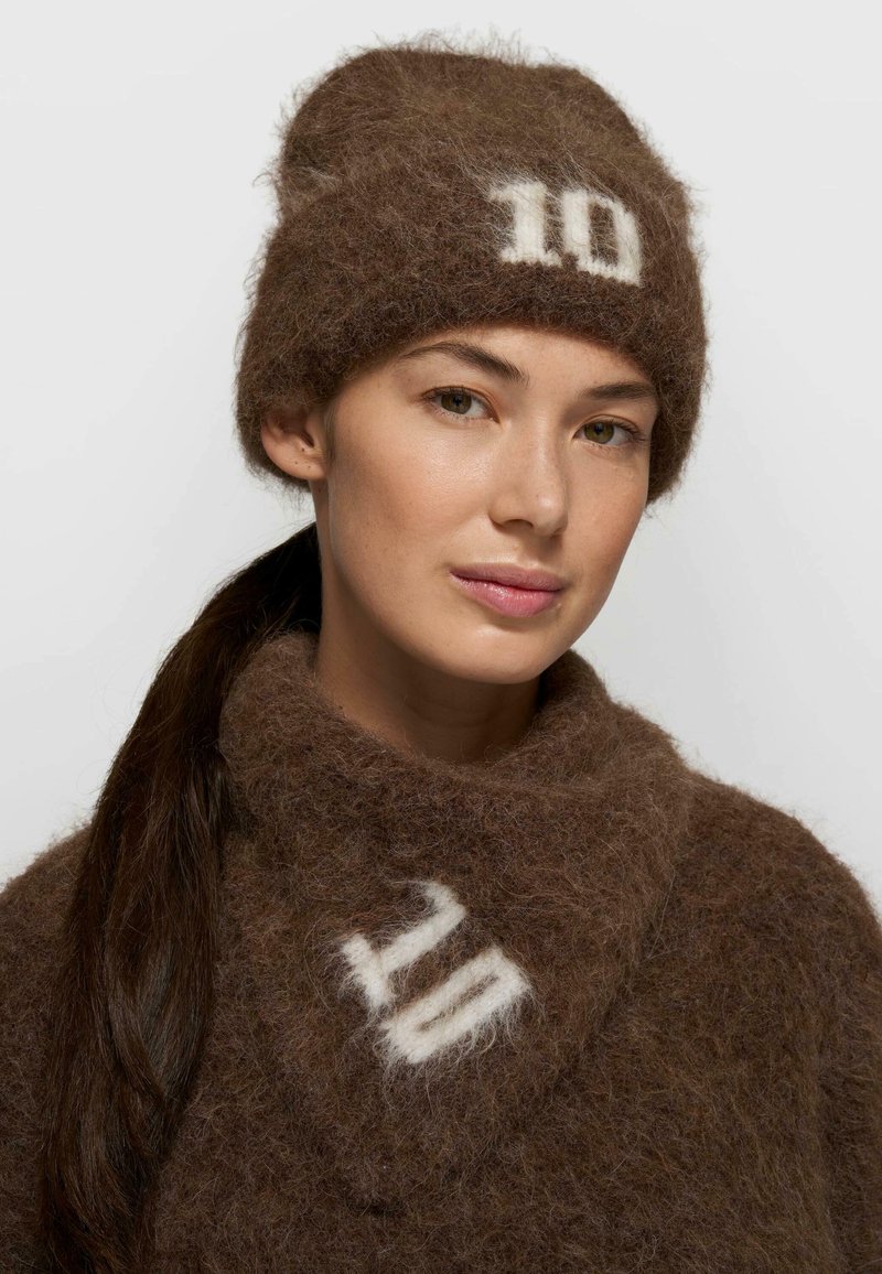 Joven con un gorro marrón peludo con el número "10" y una bufanda a juego con el detalle blanco "10" sobre un fondo claro.