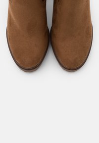 Brązowe botki zamszowe o gładkiej fakturze, zaokrąglonym nosku i lekko podniesionej gumowej podeszwie. Brak widocznych elementów metalowych ani wzorów.