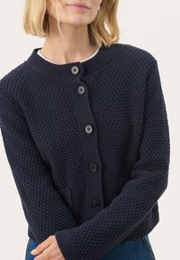 Tummansininen neulecardigan, jossa on teksturoitu kuvio, pyöreä pääntie, kuusi mustaa nappia ja sivutaskut. Lyhyt malli, jossa on pitkät hihat.