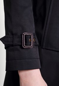 Svart ärmspets med justerbart band, utrustad med en rektangulär spänne och en mässingsöggla, tillverkad av ett mjukt, lätt tyg.