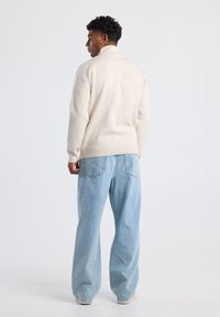 Beige knit sweater with ribbed cuffs and hem, paired with light blue wide-leg jeans. The model stands facing away against a plain background.