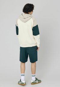 Sweat à capuche crème avec des accents vert foncé et beige, associé à un short vert foncé. Tissu doux, coupe décontractée, détails de poche visibles.