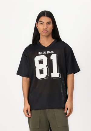 T-shirt en jersey noir à mailles avec des manches courtes, comportant un "81" blanc et le texte "GUESS JEANS". Conception à col en V, coupe décontractée et texture respirante.