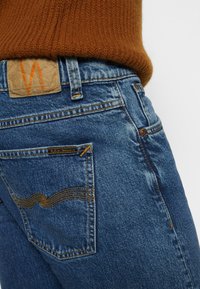 Blå denimjeans med en texturerad yta, med en patch med en orange 'W'-logotyp, synliga sömmar och en ficka med ett vågmönster.