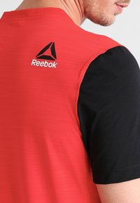 Camiseta atlética roja con mangas cortas negras, con un patrón texturizado. Logo de Reebok destacado en la parte de atrás en negro y blanco.