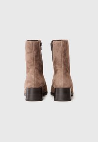 Botine din suede maro până la gleznă, cu toc stratificat, închidere cu fermoar pe lateral și o textură netedă. Prezintă un vârf rotunjit și un design elegant.