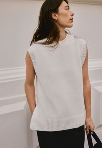 Pull sans manches en maille blanche avec encolure et ourlet côtelés, présentant une coupe décontractée et une texture douce, porté sur une jupe foncée.