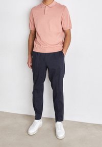 Uomo in piedi che indossa una polo rosa a maniche corte, pantaloni blu scuro con pieghe e sneakers bianche su uno sfondo bianco semplice.