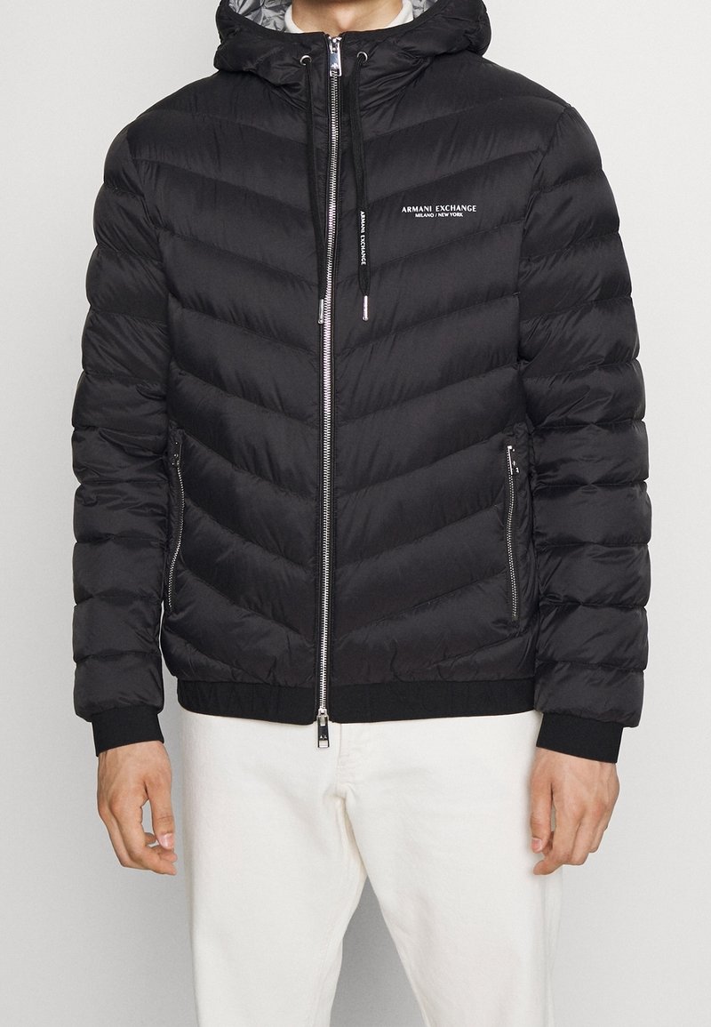Homme portant une veste matelassée noire à capuche avec zip et logo "Armani Exchange Milano/New York," associée à un pantalon blanc.