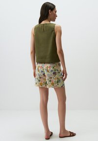 Top senza maniche in lino verde con aperture a chiave sul retro, abbinato a pantaloni corti con fantasie floreali e fogliari in più colori.