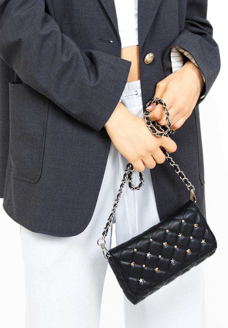Borsa a tracolla in pelle nera trapuntata con borchie a forma di stella e cuore dorate, dotata di una tracolla in catena argentata. Abbinata a un blazer scuro e pantaloni chiari.