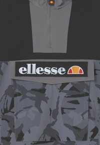 Veste pull grise et noire avec un col zippé mi-haut, présentant un motif camouflage et un logo "ellesse" brodé sur une patch rectangulaire.