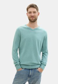 Pulover s V-izrezom v svetlo teal barvi, narejen iz mehke tkanine, z rebrastimi manšetami in pasom, v kombinaciji z modrimi kavbojkami in rokami v žepih.