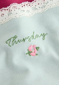 Hele sinine kangas, millel on rohelise tikandiga sõna "Neljapäev" koos roosa lillemotiiviga, on accentitud valge pitsuseelikuga ülaosas.