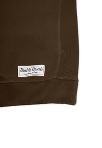 Ruskea huppari, jossa on ribbireunat hihansuissa ja helmassa. Varustettu valkoisella suorakulmaisella tarralla, jossa on teksti "Band of Rascals" ja orgaanista puuvillaa koskevia tietoja.