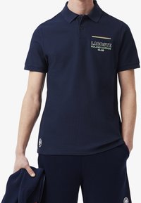 Marinblå pikétröja i texturerat tyg, med krage, två knappar på placket och broderad logotyp med färgglada accenter på bröstet.