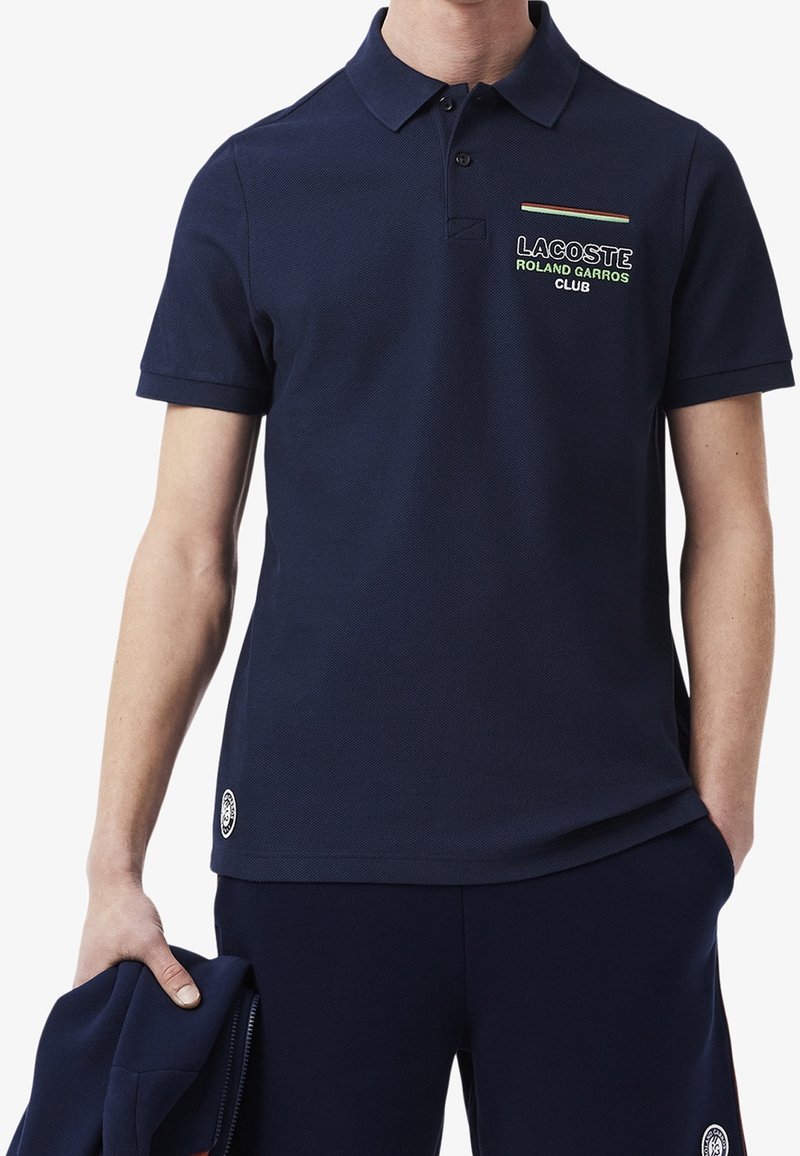 Marinblå pikétröja i texturerat tyg, med krage, två knappar på placket och broderad logotyp med färgglada accenter på bröstet.