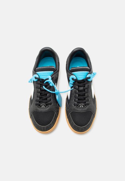 Ανδρικά αθλητικά παπούτσια ασπρόμαυρα με λαστιχένιες σόλες και φωτεινά μπλε εσωτερικά, συνδεδεμένα με μπλε πλαστική ετικέτα και κορδόνι πάνω σε λευκό φόντο.