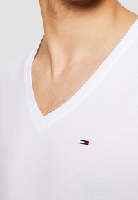 Bílý tričko s výstřihem do V vyrobené z bavlny, s malým vyšitým logem v červené, bílé a modré barvě na hrudi. Hladká textura a přiléhavý střih.