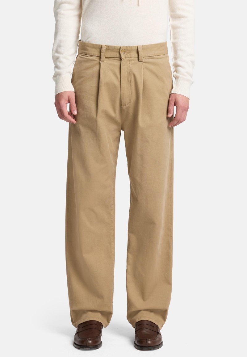 Pantaloni in cotone beige con vestibilità rilassata, dotati di pieghe frontali, chiusura a bottone e tasche laterali. Indossati con scarpe in pelle marroni.