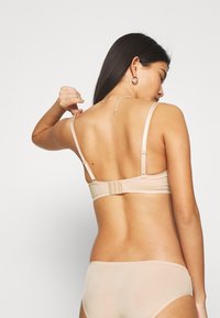 Beige wireless bra with thin adjustable straps and a hook-and-eye closure. Smooth texture and seamless design, suitable for layering.