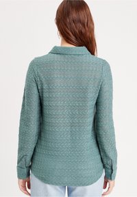 Blouse turquoise à manches longues avec un motif en zigzag texturé, col et poignets boutonnés. Le tissu semble doux et léger.