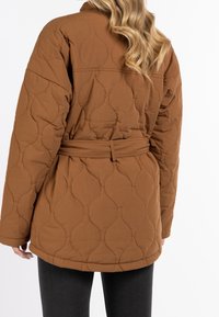 Veste matelassée marron avec une ceinture à la taille, présentant des motifs de couture courbés et une coupe décontractée. Manches longues et col haut complètent le design.