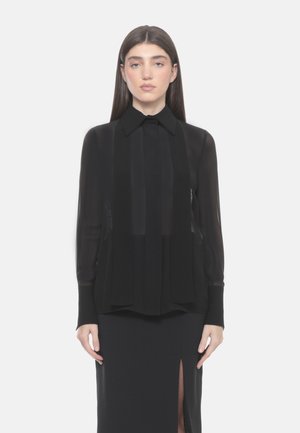 Donna con capelli scuri e lisci che indossa una camicetta nera trasparente a maniche lunghe con colletto e una gonna nera con una spacco frontale, in piedi su sfondo bianco.