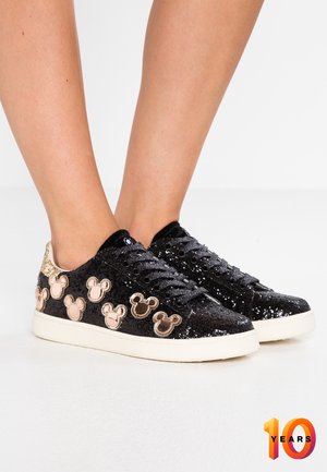 Czarne brokatowe sneakersy z brązowymi naszywkami w kształcie głowy myszy, noszone na bosych stopach na jednolitym białym tle.