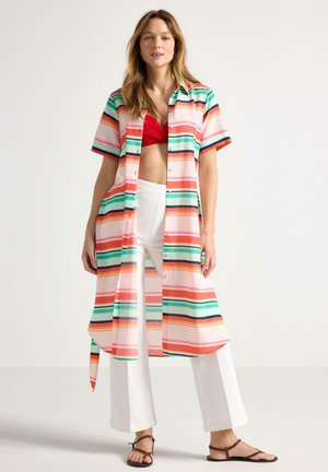 Kvinde iført lang, flerfarvet, stribet åben skjorte over rød cropped top, hvide vidde bukser og brune sandaler, stående foran en ensfarvet baggrund.