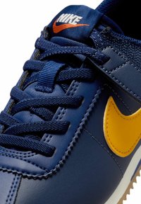 Baskets bleu foncé avec un swoosh Nike jaune. Présente un cuir texturé, des lacets contrastants et une languette rembourrée. Semelle de couleur claire.
