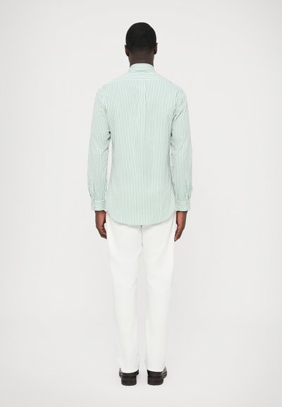 Zeleni srajca s črtami in gumbi, z dolgimi rokavi, prikazana od zadaj, kombinirana z belimi hlačami in temnimi čevlji na nevtralnem ozadju.