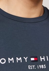 Granatowa bawełniana koszulka z okrągłym dekoltem, teksturowanym materiałem oraz haftowanym logo "TOMMY HILFIGER" w czerwonych, białych i niebieskich akcentach.