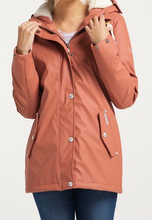 Femme portant un imperméable couleur rouille avec une capuche doublée de polaire blanche, tenant les cordons de la capuche, et portant un jean bleu.