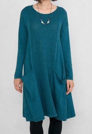 Femme portant une robe en maille manches longues de couleur bleu sarcelle avec des poches et un collier rond de perles colorées, debout devant un fond clair uni.