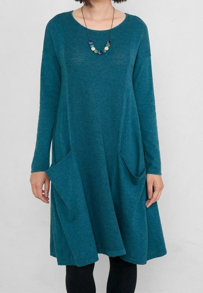 Femme portant une robe en maille manches longues de couleur bleu sarcelle avec des poches et un collier rond de perles colorées, debout devant un fond clair uni.