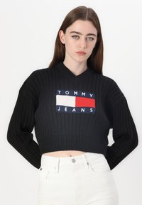 Černý žebrovaný zkrácený svetr s bílým, červeným a modrým logem "TOMMY JEANS" na hrudi, v kombinaci s bílými kalhotami.