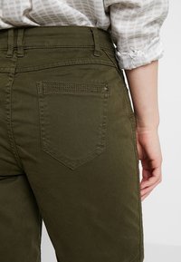 Pantalons kaki avec taille haute, grande poche arrière avec détails de couture et texture lisse. Le haut est en tissu à carreaux léger.