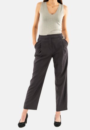 Pantalones grises a medida con pliegues, cintura alta y un emblema dorado. Combinados con una blusa sin mangas de color verde claro y tacones negros.