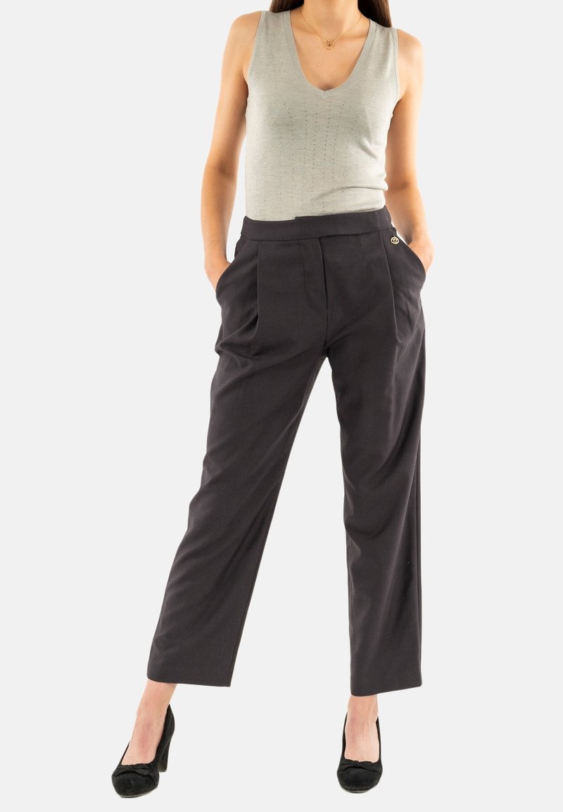 Pantalones grises a medida con pliegues, cintura alta y un emblema dorado. Combinados con una blusa sin mangas de color verde claro y tacones negros.