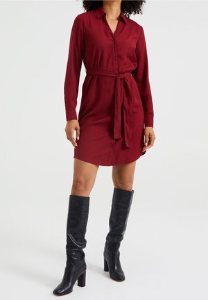 Kobieta ubrana w bordową sukienkę koszulową o długości do kolan z długimi rękawami, zapinaną na guziki, z wiązanym pasem w talii oraz czarne kozaki na wysokim obcasie do kolan, stoi.