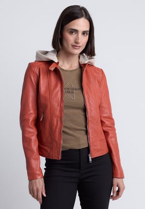 Femme aux cheveux raides et foncés portant une chemise marron ornée d'étoiles cloutées et une veste en cuir orange ouverte avec un pantalon noir sur un fond uni.