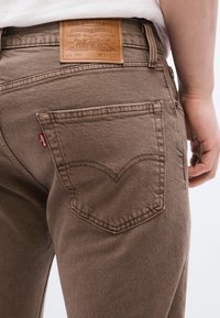 Οπίσθια όψη ατόμου που φορά καφέ τζιν Levi's, εμφανίζοντας την τσέπη, το δερμάτινο ετικέτα της μάρκας, και το χέρι που ακουμπά δίπλα στο ισχίο.