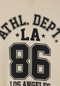 Felpa color crema con grande testo nero: "ATHL. DEPT. LA" sopra "86" e "LOS ANGELES", con accenti di stelle prominenti. Tessuto morbido.